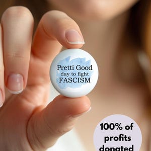 May include: A white button with the text "Pretti Good day to fight FASCISM" in black font, with a blue watercolor design. The button is held in a person's hand. A purple circle with the text "100% of profits donated" is in the lower right.