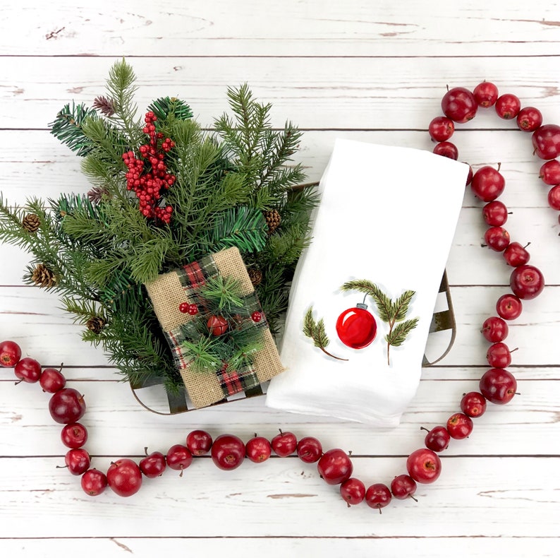 May include: A white kitchen towel with a red Christmas ornament and green pine branches embroidered on it. The towel is surrounded by a garland of red apples and a small gift wrapped in burlap with a plaid pattern.