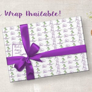 K&ouml;nnte beinhalten: Ein verpacktes Geschenk mit wei&szlig;em Papier mit gr&uuml;nen Figuren und lila Herzen. Ein breites lila Band ist um das Geschenk gebunden. Der Text "Gift Wrap Available!" steht dar&uuml;ber.