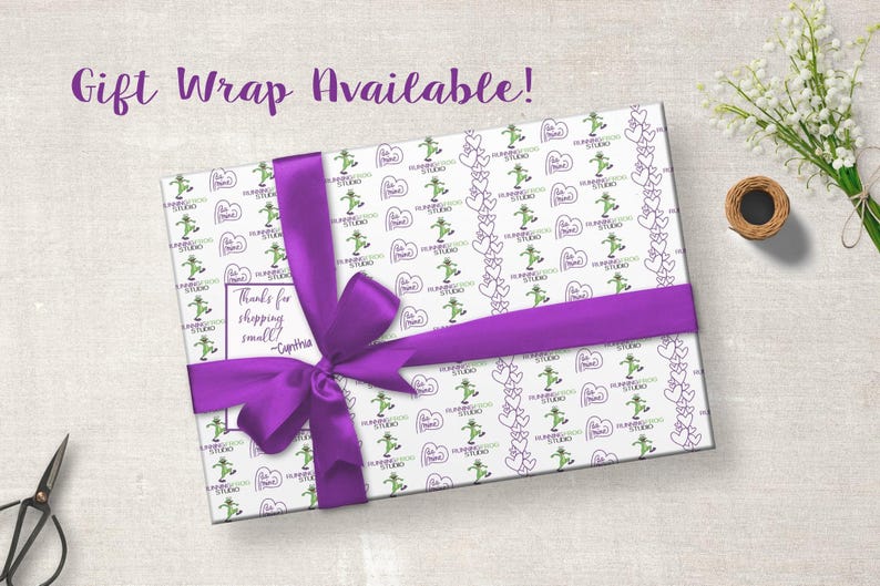 K&ouml;nnte beinhalten: Ein verpacktes Geschenk mit wei&szlig;em Papier mit gr&uuml;nen Figuren und lila Herzen. Ein breites lila Band ist um das Geschenk gebunden. Der Text "Gift Wrap Available!" steht dar&uuml;ber.