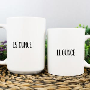 K&ouml;nnte beinhalten: Zwei wei&szlig;e Keramikbecher, einer mit der Aufschrift "15 OUNCE" und der andere mit "11 OUNCE". Die Becher haben einen gebogenen Griff und stehen vor einem neutralen Hintergrund mit Gr&uuml;n und lila Blumen. Die Becher stehen auf einer gewebten Oberfl&auml;che.
