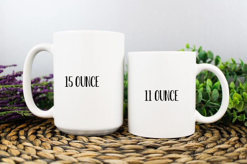 K&ouml;nnte beinhalten: Zwei wei&szlig;e Keramikbecher, einer mit der Aufschrift "15 OUNCE" und der andere mit "11 OUNCE". Die Becher haben einen gebogenen Griff und stehen vor einem neutralen Hintergrund mit Gr&uuml;n und lila Blumen. Die Becher stehen auf einer gewebten Oberfl&auml;che.