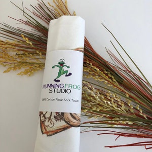 May include: A white flour sack towel with a green frog logo that says "Running Frog Studio" and "20% Cotton Flour Sack Towel". The towel is rolled up and has a brown and white floral design on the bottom.