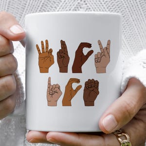 K&ouml;nnte beinhalten: Wei&szlig;er Keramikbecher mit Illustrationen von H&auml;nden, die das Wort "LOVE" in amerikanischer Geb&auml;rdensprache buchstabieren. Die H&auml;nde sind in verschiedenen Hautt&ouml;nen dargestellt, mit einem einfachen Design. Der Becher wird von einer Person gehalten.