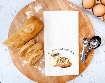 May include: A white kitchen towel with the text "in my sourdough era" and a watercolor illustration of a jar and loaf of bread. A sliced baguette, measuring spoons, and a carton of eggs are also present on a wooden cutting board.