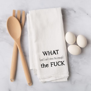 May include: White kitchen towel with the text "WHAT (and I can't stress this enough) the FUCK" in black. A wooden spoon and fork are on the left, and three white eggs are on the right.