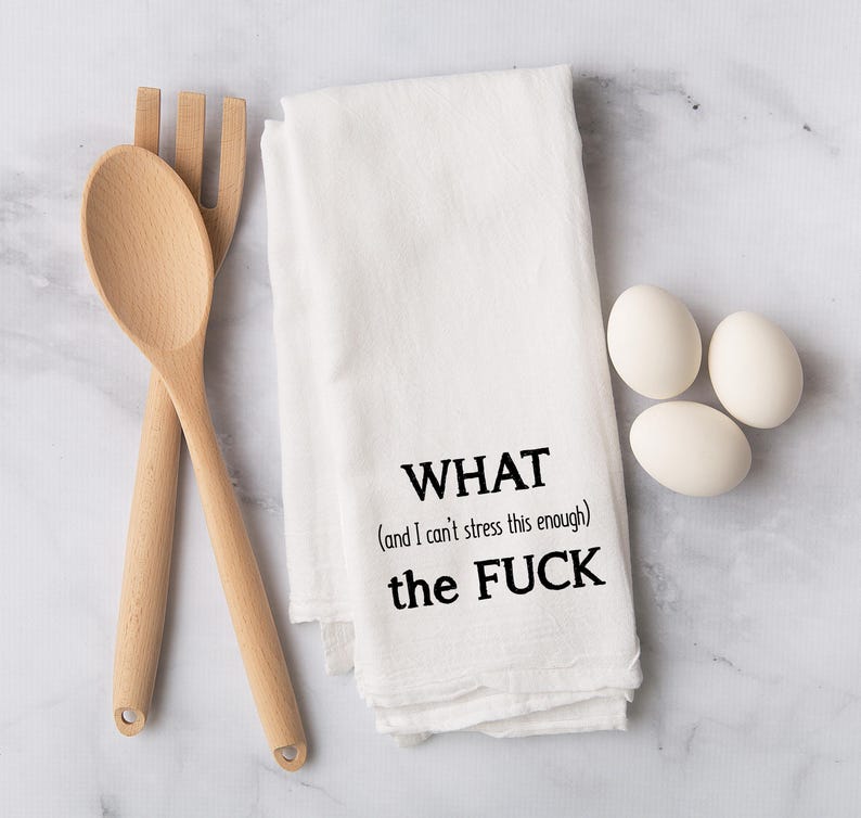 May include: White kitchen towel with the text "WHAT (and I can't stress this enough) the FUCK" in black. A wooden spoon and fork are on the left, and three white eggs are on the right.