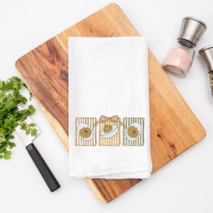 May include: White kitchen towel with an Easter-themed design featuring three illustrated squares with eggs and bows. The towel is placed on a wooden cutting board, with a bunch of cilantro, a knife, and salt and pepper shakers nearby.