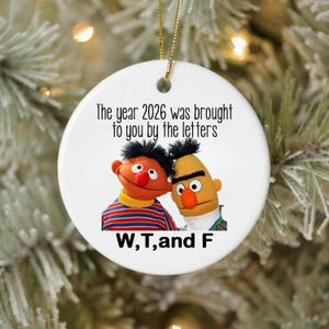 May include: A round white ceramic ornament featuring an illustration of two puppets, one orange with a striped shirt and one yellow with a black hair tuft. The text reads: The year 2026 was brought to you by the letters W, T, and F.