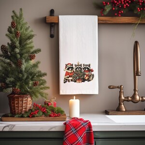 K&ouml;nnte beinhalten: Wei&szlig;es K&uuml;chentuch mit einem weihnachtlichen Design von drei Waschb&auml;ren, die sich um einen geschm&uuml;ckten Baum versammelt haben. Ein kleiner Weihnachtsbaum und eine Kerze sind im Vordergrund zu sehen.