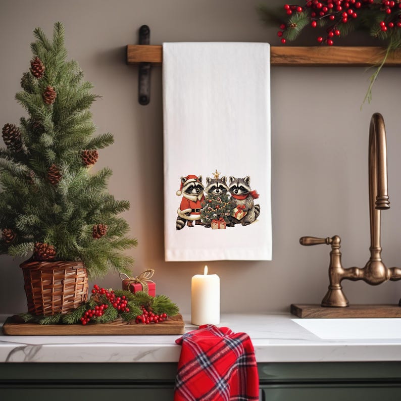 K&ouml;nnte beinhalten: Wei&szlig;es K&uuml;chentuch mit einem weihnachtlichen Design von drei Waschb&auml;ren, die sich um einen geschm&uuml;ckten Baum versammelt haben. Ein kleiner Weihnachtsbaum und eine Kerze sind im Vordergrund zu sehen.