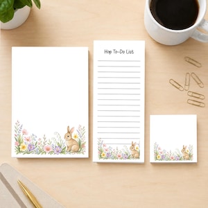 May include: A set of three white paper notepads featuring watercolor illustrations of a brown rabbit surrounded by colorful spring flowers. The tall, narrow notepad includes horizontal lines and the printed text Hop-To-Do List at the top.