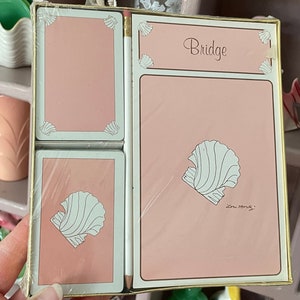 May include: A vintage bridge card game set in a pink and gold box. The cards and score pad feature a white seashell design. The box is held in a hand, with a shelf of decorative items in the background.