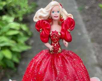 May include: A Christmas ornament featuring a doll in a red gown with gold glitter accents. The doll has blonde hair, red earrings, and a red bodice. The ornament is held in a hand.