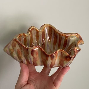 May include: A ceramic seashell-shaped bowl with a glossy finish. The bowl features a wavy, textured design with shades of brown, tan, and hints of purple. The bowl is held in a hand against a plain background.