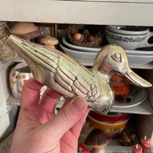 May include: A silver-colored duck figurine with detailed wing and feather patterns. The duck has a long beak and is held in a hand. The background includes shelves with various decorative items, including a ceramic bowl and teacups.