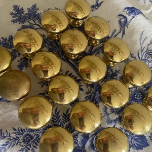 May include: A collection of shiny, gold-colored cabinet knobs with a rounded top and a short stem. The knobs are arranged on a white cloth with a blue floral pattern. The knobs are likely made of metal.
