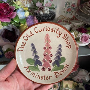 May include: A small, round ceramic plate with a cream-colored background and a brown rim. The plate features a floral design with pink and purple flowers and green leaves. The text "The Old Curiosity Shop Axminster Devon" is written in brown script around the edge.