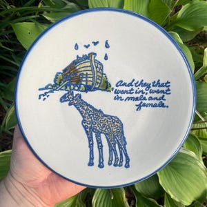 May include: A white ceramic plate with a blue rim and a blue illustration of Noah's Ark, rain, and two giraffes. The plate has the text "And they that went in, went in male and female."