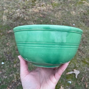 May include: A vintage, green ceramic bowl with horizontal ridged detailing. The bowl is held in a hand, showcasing its rounded shape and glossy finish. The bowl's color is a vibrant, solid green.