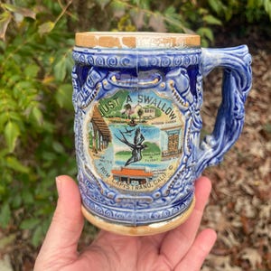May include: Blue ceramic beer stein with a handle, featuring a detailed relief design and the words "Just a Swallow" with images of a mission and swallows. The stein has a wooden lid and is from Capistrano, California.