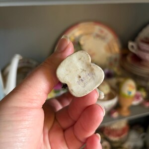 May include: A small, off-white ceramic figurine in the shape of a heart. The bottom of the figurine has the text "MADE IN JAPAN" stamped in purple ink. The figurine is held in a hand, with other decorative items in the background.