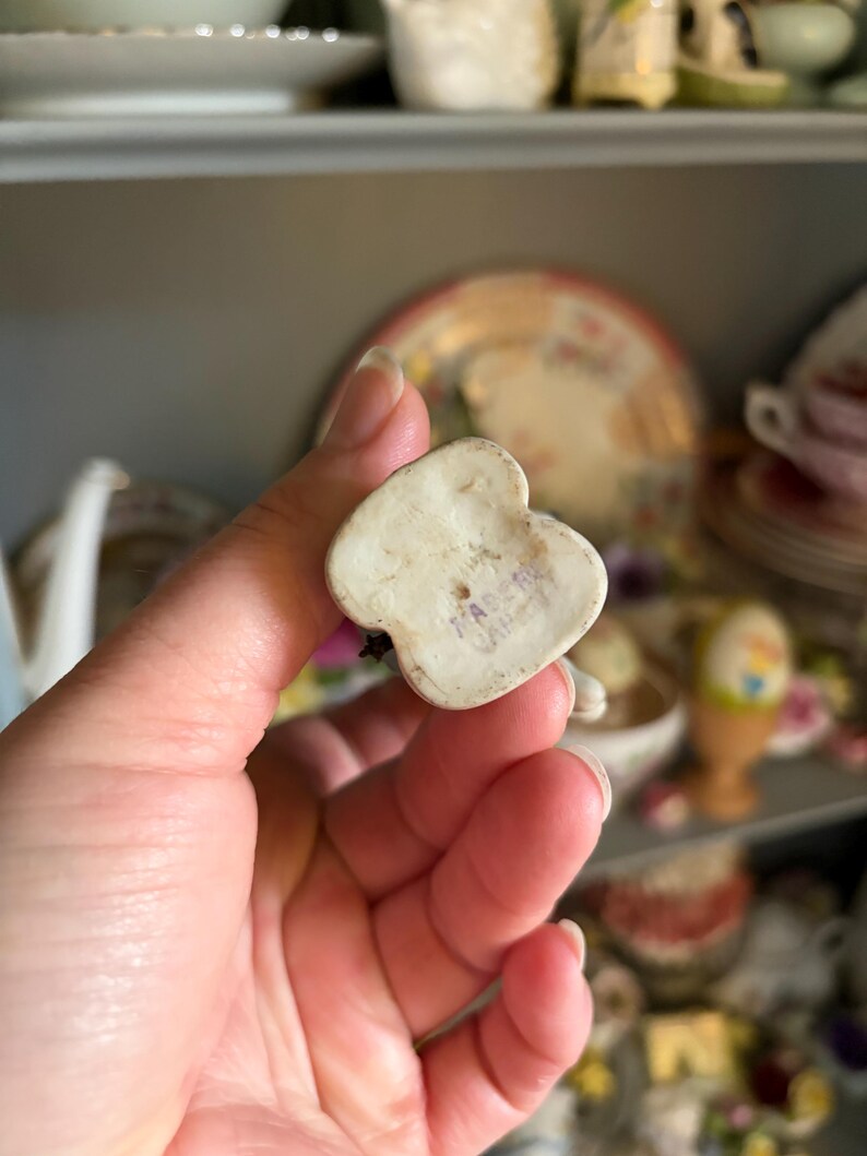 May include: A small, off-white ceramic figurine in the shape of a heart. The bottom of the figurine has the text "MADE IN JAPAN" stamped in purple ink. The figurine is held in a hand, with other decorative items in the background.