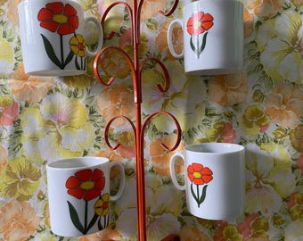 May include: An orange metal mug tree holds four white ceramic mugs. Each mug features a red and yellow flower design with green stems. The mug tree has a decorative, curly design and is set against a floral patterned background.
