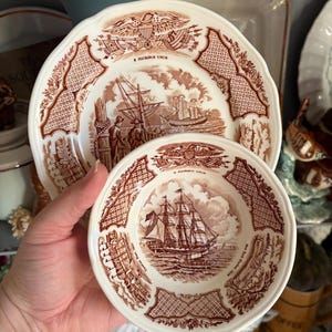 May include: A cream-colored ceramic plate and a matching smaller bowl, both featuring brown transferware illustrations of sailing ships, the text E PLURIBUS UNUM, and decorative borders. The bowl includes the text Canton Port 1850.