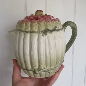May include: A cream-colored ceramic teapot with a lid. The lid and handle are green, and the top of the lid is a reddish-pink color. The teapot has vertical ridges and a decorative rope-like design around the middle.