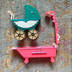 May include: A teal and white plastic doll carriage with cream-colored wheels and a separate bright pink plastic doll bathtub featuring a tall, floral-detailed shower attachment with a pink shower head and small purple accents on the feet.