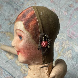 May include: A vintage doll head with painted features, including red hair, rosy cheeks, and red lips. The doll wears a green head covering with a small red flower accent. The doll's arm is visible, and the doll is made of fabric.