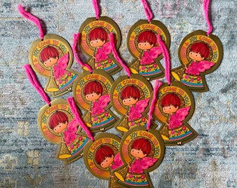 Puede incluir: Un conjunto de adornos de ángeles vintage. Cada adorno dorado presenta un ángel de dibujos animados con cabello rojo y alas rosas, sosteniendo una trompeta. El texto "LOVE & JOY COME TO YOU" está en un diseño circular. Están ensartados con hilo rosa.