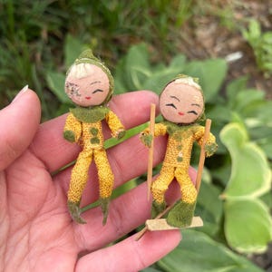 May include: Two small, vintage elf dolls. Each doll is dressed in yellow and green fabric with a smiling face. One doll is standing, and the other is on skis made of wood and cardboard. The dolls are held in a hand against a background of green foliage.