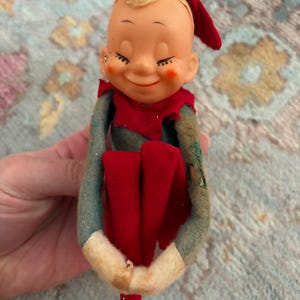 May include: A vintage Christmas elf doll with a smiling face, rosy cheeks, and closed eyes. The elf wears a red hat, collar, and pants, with a green and blue body and white cuffs. The doll is a nostalgic holiday decoration.