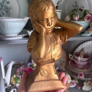 May include: A gold-colored bust of a child covering their ears with their hands. The sculpture has a matte finish and is set against a backdrop of vintage dishware. The bust is on a pedestal with a snail design.
