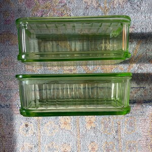 May include: Two rectangular, light green glass dishes. Each dish has a ribbed interior and a slightly raised rim. The dishes are positioned on a patterned surface, with one slightly above the other.