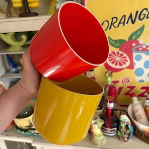 May include: Two cylindrical containers, one red and one yellow, are held up. The red container is on top, and the yellow one is below. Both have a glossy finish and a white rim. The background includes a vintage "ORANGE" sign.