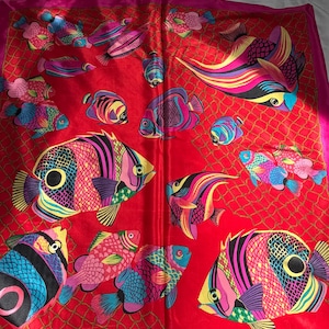 May include: A vibrant red square scarf featuring a colorful fish pattern. The fish are depicted in various sizes and orientations, with a mix of bright colors including pink, blue, yellow, and purple. The scarf has a pink border and a net-like design.