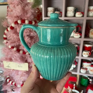 May include: A teal blue ceramic creamer with a lid and ribbed design. The creamer is being held in a hand.