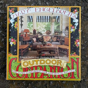 May include: Book cover with a colorful illustration of a porch with wicker furniture and a floral design border. The title is "Mary Engelbreit's Outdoor Companion: The Mary Engelbreit Look and How to Get It!"