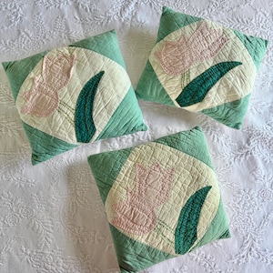 May include: Three square decorative pillows with a floral design. Each pillow features a light green quilted border, a cream-colored center, and a pink tulip with a green leaf. The pillows are arranged on a white, patterned surface.