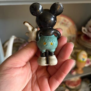 May include: A vintage Mickey Mouse figurine, viewed from the back. The figure has black ears and head, white gloves, and light blue shorts. The words "Mickey Mouse" are printed on the shorts. The figurine is standing on white shoes.