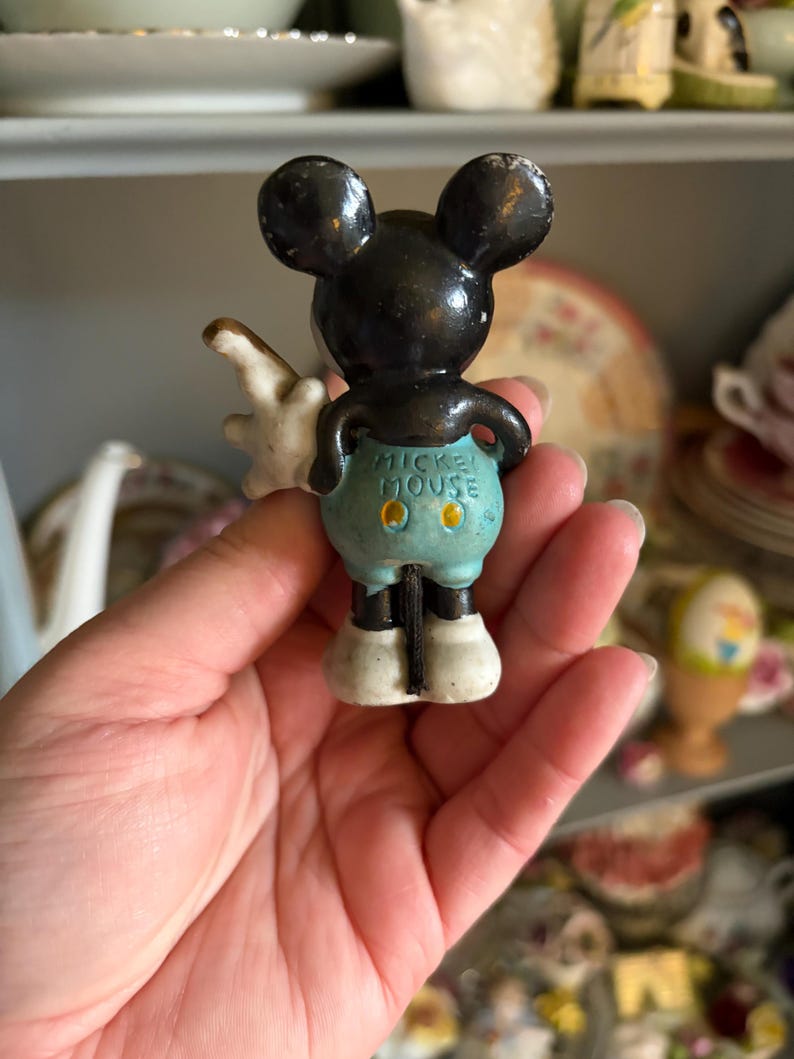 May include: A vintage Mickey Mouse figurine, viewed from the back. The figure has black ears and head, white gloves, and light blue shorts. The words "Mickey Mouse" are printed on the shorts. The figurine is standing on white shoes.