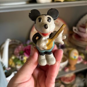 May include: A vintage Mickey Mouse figurine, featuring black ears, a white face, and a red mouth. The figure is holding a yellow banjo and wearing blue shorts. The figurine is a collectible item.