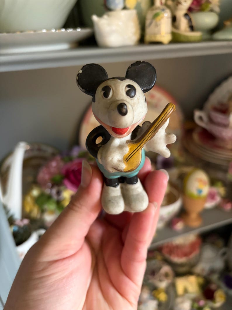 May include: A vintage Mickey Mouse figurine, featuring black ears, a white face, and a red mouth. The figure is holding a yellow banjo and wearing blue shorts. The figurine is a collectible item.