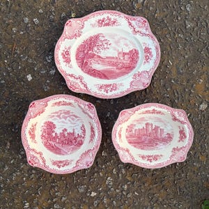 May include: Three antique white and pink ceramic serving dishes. The dishes feature a detailed pink landscape scene with a castle and floral border. The set includes a large oval platter and two smaller dishes.