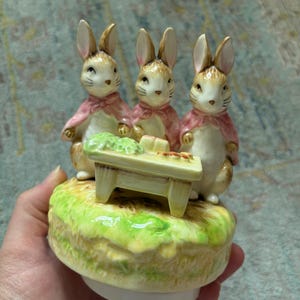 May include: A ceramic music box figurine featuring three brown rabbits with pink capes standing around a small yellow table. The rabbits are positioned on a green grassy base. The table holds small green and orange items. The base is round and white.