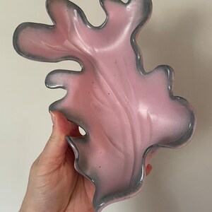 May include: A pink, leaf-shaped decorative dish with a gray trim. The dish has a smooth, glossy finish and a unique, organic design. The dish is held in a hand against a white background. This decorative dish is ideal for holding small items.