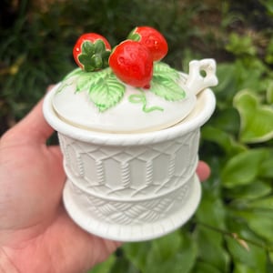 May include: A cream-colored ceramic sugar bowl with a textured basket-weave pattern on the base. The lid features three sculpted red strawberries with green leaves and a small handle. The bowl has a matching lid with a cutout for a spoon.